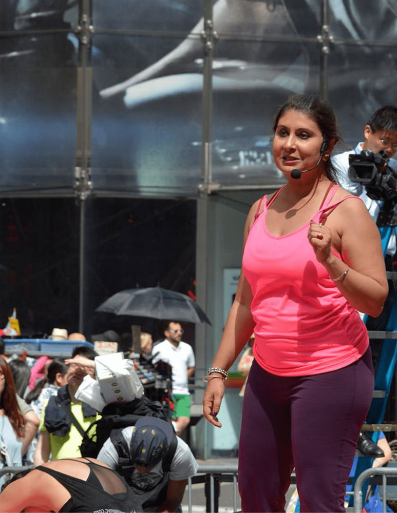 Rajashree leading an outdoor yoga class in NYC, wearing a headset and bright activewear, with a crowd and camera crew nearby